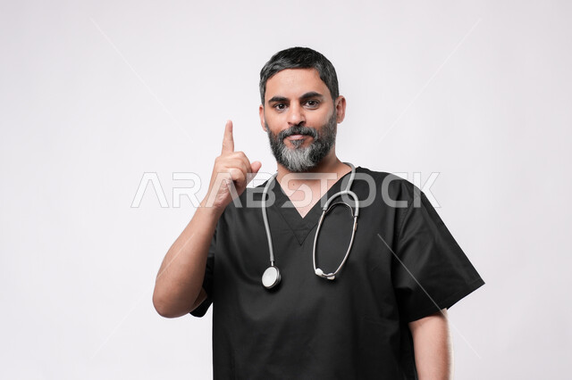 الاستعداد لتقديم الخدمات للمرضى بكل كفاءه، مهن ووظائف طبية في القطاع الصحي, مفهوم الطب والرعاية الصحية، بورتريه لطبيب كبير بالسن عربي خليجي يرتدي اليونيفورم وسماعه الفحص يرفع اصبع السبابة للاعلى للاشارة الى شيء ما, خلفية بيضاء