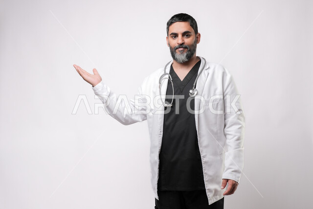 الترحيب والاستقبال ببشاشة، بورتريه لطبيب مسن عربي سعودي خليجي يرتدي يرتدي المعطف الطبي والسماعه ينظر الى الكاميرا يفتح كفه بايماءات السرور برؤية شخص ما، الاستعداد لتقديم الخدمات للمرضى بكل كفاءه، مفهوم الطب والرعاية الصحية، الإشارة نحو هدف معين، خلفية بيضاء