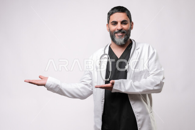 الترحيب والاستقبال ببشاشة، بورتريه لطبيب مسن عربي سعودي خليجي يرتدي يرتدي المعطف الطبي والسماعه ينظر الى الكاميرا يفتح كفيه بايماءات السرور برؤية شخص ما، الاستعداد لتقديم الخدمات للمرضى بكل كفاءه، مفهوم الطب والرعاية الصحية، الإشارة نحو هدف معين، خلفية بيضاء