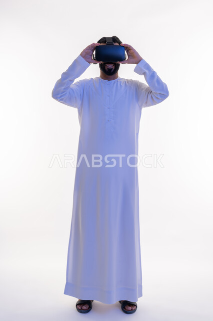 التفاعل مع الفيلم ومحاكاة العالم الخيالي، بورتريه لرجل عربي خليجي اماراتي مبتسم يرتدي الكندورة التقليدية يقوم بمشاهدة أفلام عبر نظارات الواقع الافتراضي ثلاثية الأبعاد, ايماءات تدل على الاندماج والمتعة, صورة الجسم بالطول الكامل، خلفيه بيضاء