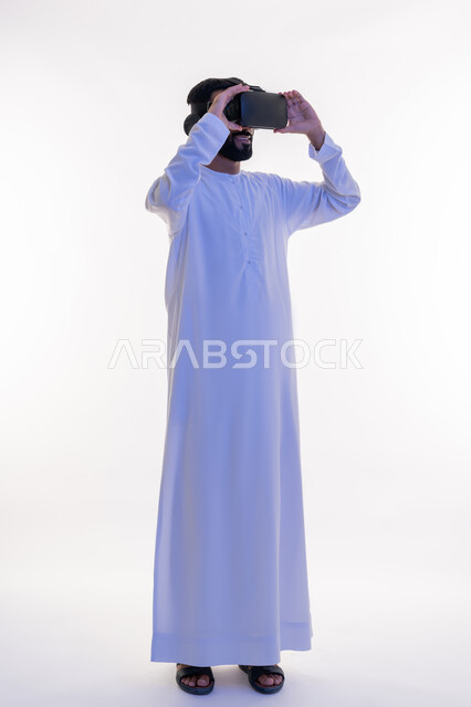 التفاعل مع الفيلم ومحاكاة العالم الخيالي، بورتريه لرجل عربي خليجي اماراتي مبتسم يرتدي الكندورة التقليدية يقوم بمشاهدة أفلام عبر نظارات الواقع الافتراضي ثلاثية الأبعاد, ايماءات تدل على الاندماج والمتعة, صورة الجسم بالطول الكامل، خلفيه بيضاء