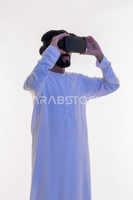 التفاعل مع الفيلم ومحاكاة العالم الخيالي، بورتريه لرجل عربي خليجي اماراتي مبتسم يرتدي الكندورة التقليدية يقوم بمشاهدة أفلام عبر نظارات الواقع الافتراضي ثلاثية الأبعاد, ايماءات تدل على الاندماج والمتعة, خلفيه بيضاء