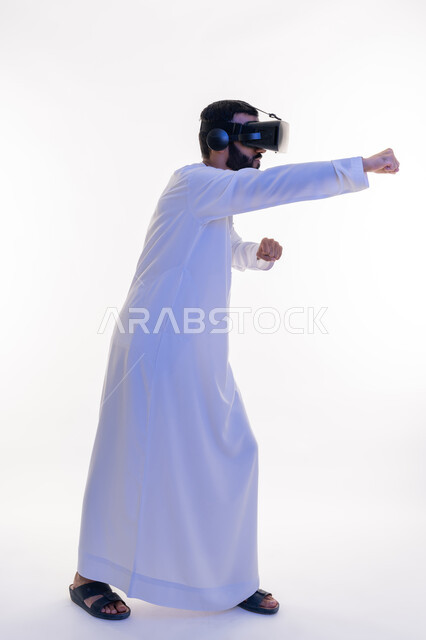 تفريغ الطاقة السلبية، التدرب على الحركات القتالية من خلال ألعاب الفيديو، ممارسة رياضة الدفاع عن النفس بتقنيات تكنولوجية، بورتريه لرجل عربي خليجي اماراتي مبتسم يرتدي الكندورة ونظارات الواقع الافتراضي بإيماءات الملاكمة والمصارعة، صورة الجسم بالطول الكامل، خلفية بيضاء