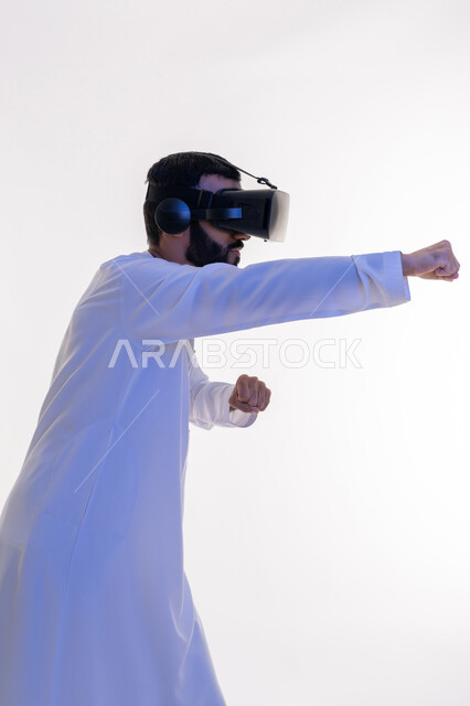 التدرب على الحركات القتالية من خلال ألعاب الفيديو، تفريغ الطاقة السلبية، ممارسة رياضة الدفاع عن النفس بتقنيات تكنولوجية، بورتريه لرجل عربي خليجي اماراتي مبتسم يرتدي الكندورة ونظارات الواقع الافتراضي بإيماءات الملاكمة والمصارعة، خلفية بيضاء