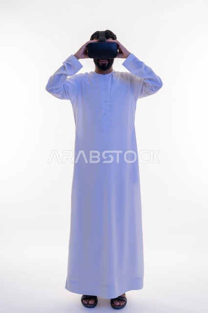 التفاعل مع الفيلم ومحاكاة العالم الخيالي، بورتريه لرجل عربي خليجي اماراتي مبتسم يرتدي الكندورة التقليدية يقوم بمشاهدة أفلام عبر نظارات الواقع الافتراضي ثلاثية الأبعاد, ايماءات تدل على الاندماج والمتعة, صورة الجسم بالطول الكامل، خلفيه بيضاء