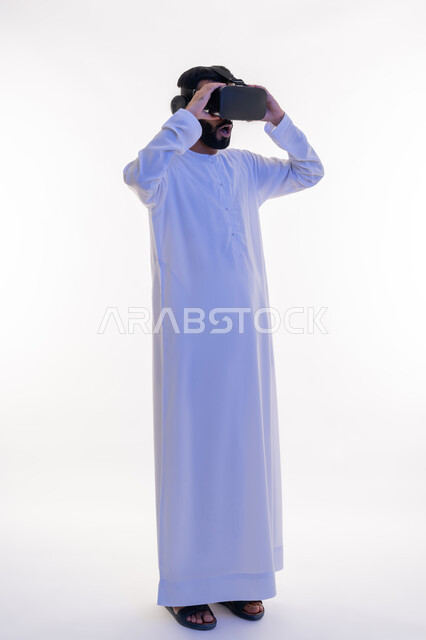 التفاعل مع الفيلم ومحاكاة العالم الخيالي، بورتريه لرجل عربي خليجي اماراتي مبتسم يرتدي الكندورة التقليدية يقوم بمشاهدة أفلام عبر نظارات الواقع الافتراضي ثلاثية الأبعاد, ايماءات تدل على الاندماج والمتعة, صورة الجسم بالطول الكامل، خلفيه بيضاء