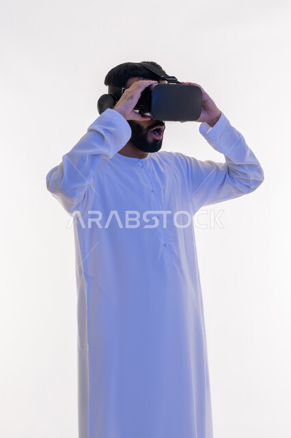 الاندهاش والاستغراب والتفاعل مع الفيلم ومحاكاة العالم الخيالي، بورتريه لرجل عربي خليجي اماراتي مبتسم يرتدي الكندورة التقليدية يقوم بمشاهدة أفلام عبر نظارات الواقع الافتراضي ثلاثية الأبعاد, ايماءات تدل على الاندماج والمتعة, خلفيه بيضاء