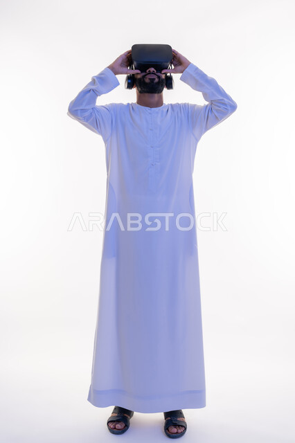 التفاعل مع الفيلم ومحاكاة العالم الخيالي، بورتريه لرجل عربي خليجي اماراتي مبتسم يرتدي الكندورة التقليدية يقوم بمشاهدة أفلام عبر نظارات الواقع الافتراضي ثلاثية الأبعاد, ايماءات تدل على الاندماج والمتعة, صورة الجسم بالطول الكامل، خلفيه بيضاء