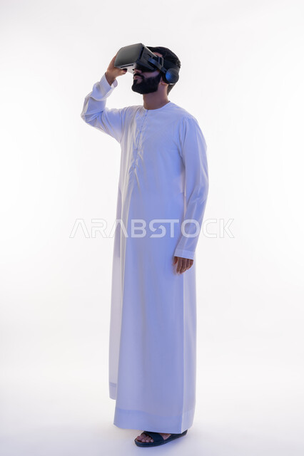 التفاعل مع الفيلم ومحاكاة العالم الخيالي، بورتريه لرجل عربي خليجي اماراتي مبتسم يرتدي الكندورة التقليدية يقوم بمشاهدة أفلام عبر نظارات الواقع الافتراضي ثلاثية الأبعاد, ايماءات تدل على الاندماج والمتعة, صورة الجسم بالطول الكامل، خلفيه بيضاء