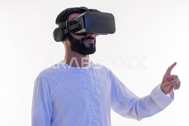 Using the sense of touch to touch holographic elements with tapping gestures, merging, interacting and simulating the imaginary world, close-up portrait of a young Saudi Emirati Arab man wearing a kandura and 3D virtual VR glasses pointing at something with his index finger, white background