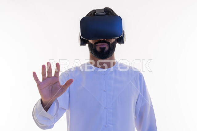 الاندماج والتفاعل و محاكاة العالم الخيالي، بورتريه مقرب لشاب عربي سعودي اماراتي يرتدي الكندورة ونظارة في آر الافتراضية ثلاثية الأبعاد يحاول الامساك بشيء ما، استخدام حاسة اللمس لملامسة عناصر هولوجرامية بإيماءات النقر, خلفية بيضاء