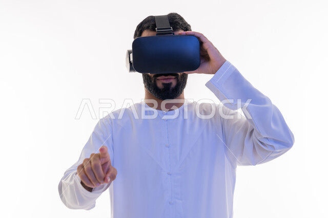 استخدام حاسة اللمس لملامسة عناصر هولوجرامية بإيماءات النقر, الاندماج والتفاعل و محاكاة العالم الخيالي، بورتريه مقرب لشاب عربي سعودي اماراتي يرتدي الكندورة ونظارة في آر الافتراضية ثلاثية الأبعاد يشير إلى شيء ما بأصبعه السبابة، خلفية بيضاء