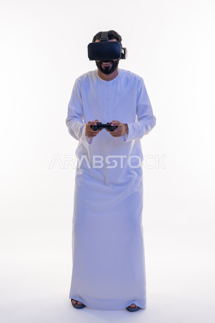أجهزة تكنولوجية وتقنيات حديثة، دمج العالم الخيالي مع المتعة والتسلية، بورتريه لرجل عربي خليجي اماراتي مبتسم يرتدي الكندورة التقليدية ونظارة الواقع الإفتراضي يمسك بيده عصا التحكم يقوم باللعب في ألعاب الفيديو  الترفيهية، صورة الجسم بالطول الكامل، خلفية بيضاء