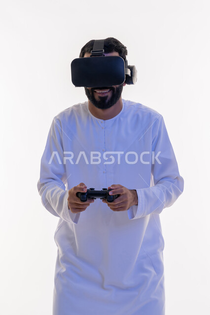 أجهزة تكنولوجية وتقنيات حديثة، دمج العالم الخيالي مع المتعة والتسلية، بورتريه لرجل عربي خليجي اماراتي مبتسم يرتدي الكندورة التقليدية ونظارة الواقع الإفتراضي يمسك بيده عصا التحكم يقوم باللعب في ألعاب الفيديو  الترفيهية، خلفية بيضاء