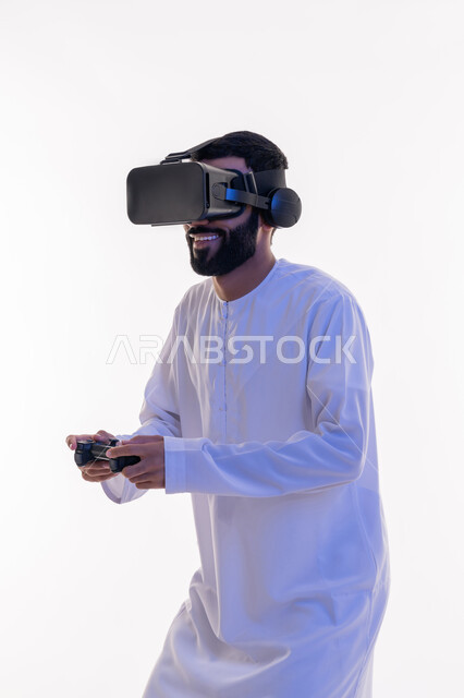اللعب في ألعاب الفيديو الترفيهية، دمج العالم الخيالي مع المتعة والتسلية، أجهزة تكنولوجية وتقنيات حديثة، بورتريه لرجل عربي خليجي اماراتي مبتسم يرتدي الكندورة التقليدية ونظارة الواقع الإفتراضي يمسك بيده عصا التحكم، خلفية بيضاء