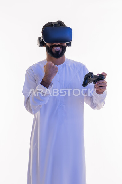 رفع قبضة اليد بايماءات الحماس، دمج العالم الخيالي مع المتعة والتسلية، أجهزة تكنولوجية وتقنيات حديثة، بورتريه لرجل عربي خليجي اماراتي مبتسم يرتدي الكندورة التقليدية ونظارة الواقع الإفتراضي يمسك بيده عصا التحكم يقوم باللعب في ألعاب الفيديو الترفيهية،  خلفية بيضاء