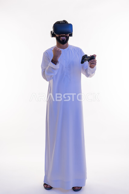اللعب في ألعاب الفيديو الترفيهية، رفع قبضة اليد بايماءات الحماس، دمج العالم الخيالي مع المتعة والتسلية، أجهزة تكنولوجية وتقنيات حديثة، بورتريه لرجل عربي خليجي اماراتي يرتدي الكندورة   ونظارة الواقع الإفتراضي يمسك بيده عصا التحكم، الجسم بالطول الكامل، خلفية بيضاء