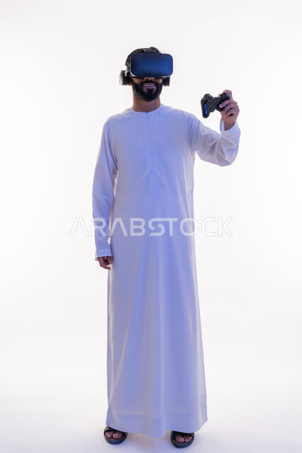أجهزة تكنولوجية وتقنيات حديثة، دمج العالم الخيالي مع المتعة والتسلية، بورتريه لرجل عربي خليجي اماراتي مبتسم يرتدي الكندورة التقليدية ونظارة الواقع الإفتراضي يمسك بيده عصا التحكم يقوم باللعب في ألعاب الفيديو الترفيهية، صورة الجسم بالطول الكامل، خلفية بيضاء