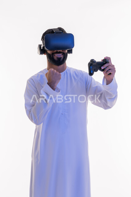 Raising fist with enthusiastic gestures, merging fantasy world with fun and entertainment, technological devices and modern techniques, portrait of a smiling Emirati Gulf Arab man wearing traditional kandura and virtual reality glasses holding joystick in his hand playing entertaining video games, white background