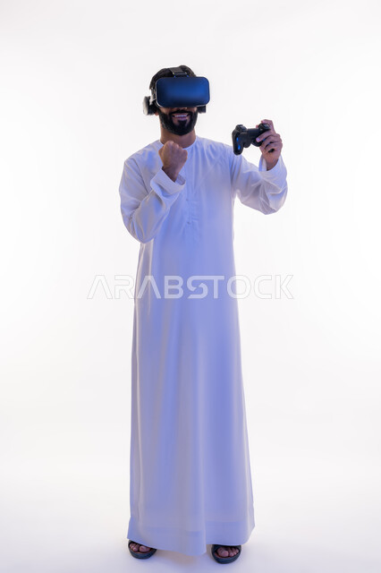 اللعب في ألعاب الفيديو الترفيهية، رفع قبضة اليد بايماءات الحماس، دمج العالم الخيالي مع المتعة والتسلية، أجهزة تكنولوجية وتقنيات حديثة، بورتريه لرجل عربي خليجي اماراتي يرتدي الكندورة   ونظارة الواقع الإفتراضي يمسك بيده عصا التحكم، الجسم بالطول الكامل، خلفية بيضاء