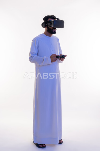 أجهزة تكنولوجية وتقنيات حديثة، دمج العالم الخيالي مع المتعة والتسلية، بورتريه لرجل عربي خليجي اماراتي مبتسم يرتدي الكندورة التقليدية ونظارة الواقع الإفتراضي يمسك بيده عصا التحكم يقوم باللعب في ألعاب الفيديو الترفيهية، صورة الجسم بالطول الكامل، خلفية بيضاء