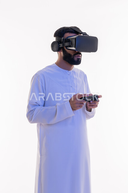 دمج العالم الخيالي مع المتعة والتسلية، أجهزة تكنولوجية وتقنيات حديثة، بورتريه لرجل عربي خليجي اماراتي مبتسم يرتدي الكندورة التقليدية ونظارة الواقع الإفتراضي يمسك بيده عصا التحكم يقوم باللعب في ألعاب الفيديو الترفيهية،  خلفية بيضاء