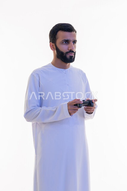 الاستمتاع بالتقنيات والتكنولوجيا الحديثة، قضاء وقت ممتع في اللعب بألعاب الفيديو، مفهوم المتعة والتسلية، إيماءات الاندماج والتفاعل,  بورتريه لرجل عربي خليجي اماراتي يرتدي الكندورة يمسك بيده عصا التحكم، خلفية بيضاء