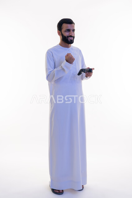 Raising the fist up with expressions of victory and triumph, gestures of enjoyment, integration and interaction, using technological devices and modern techniques, portrait of an Arab Gulf Emirati man wearing a kandura and a ghutra holding a joystick in his hand, playing recreational video games, white background