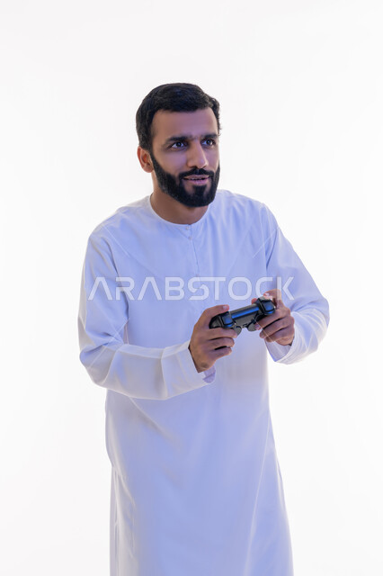 مفهوم المتعة والتسلية، إيماءات الاندماج والتفاعل, قضاء وقت ممتع في اللعب بألعاب الفيديو، الاستمتاع بالتقنيات والتكنولوجيا الحديثة، بورتريه لرجل عربي خليجي اماراتي يرتدي الكندورة يمسك بيده عصا التحكم، خلفية بيضاء
