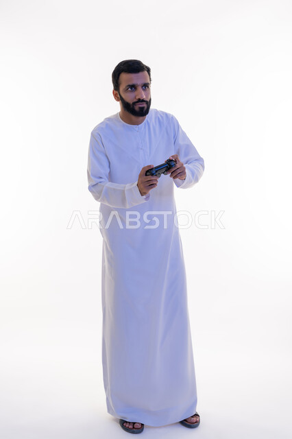 قضاء وقت ممتع في اللعب بألعاب الفيديو، الاستمتاع بالتقنيات والتكنولوجيا الحديثة، مفهوم المتعة والتسلية، إيماءات الاندماج والتفاعل,  بورتريه لرجل عربي خليجي اماراتي يرتدي الكندورة يمسك بيده عصا التحكم، الجسم بالطول الكامل، خلفية بيضاء