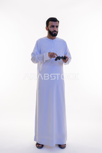 استخدام الأجهزة التكنولوجية والتقنيات الحديثة، إيماءات الاستمتاع والاندماج والتفاعل, بورتريه لرجل عربي خليجي اماراتي يرتدي الكندورة يقوم بالنقر على عصا التحكم، اللعب بألعاب الفيديو الترفيهية، الطول الكامل للجسم، خلفية بيضاء