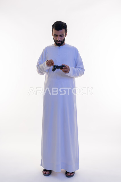 Playing recreational video games, using technological devices and modern techniques, gestures of enjoyment, integration and interaction, portrait of an Arab Gulf Emirati man wearing a kandura and a ghutra clicking on a joystick, full body length, white background