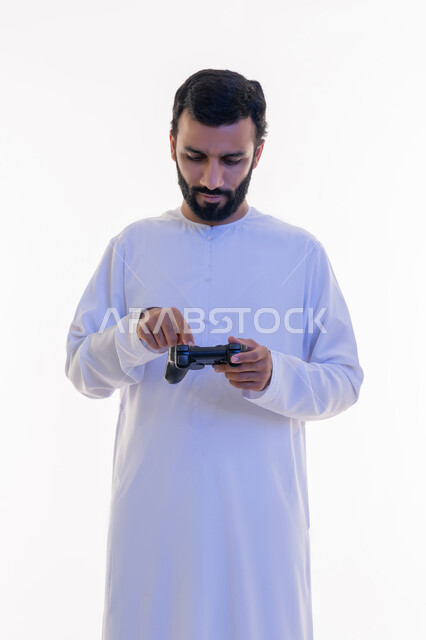 Gestures of enjoyment, integration and interaction, use of technological devices and modern techniques, portrait of an Arab Gulf Emirati man wearing a kandura and ghutra clicking on a joystick, playing recreational video games, white background