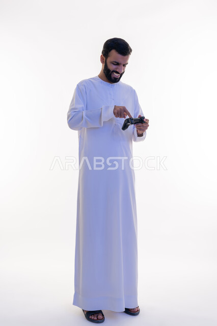 استخدام الأجهزة التكنولوجية والتقنيات الحديثة، إيماءات الاستمتاع والاندماج والتفاعل, بورتريه لرجل عربي خليجي اماراتي يرتدي الكندورة يقوم بالنقر على عصا التحكم، اللعب بألعاب الفيديو الترفيهية، الطول الكامل للجسم، خلفية بيضاء