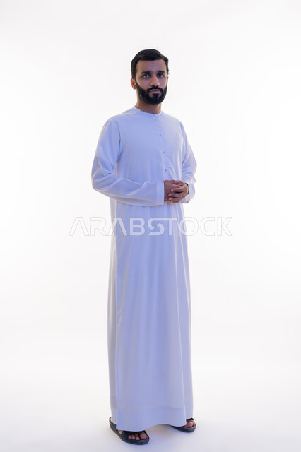 Manliness, strength and self-confidence, looking at the camera with gestures of seriousness and pleasure, interest in elegance and outward appearance, portrait of an Arab Gulf Emirati man wearing a kandura and ghutra, standing with clasped hands with expressions of firmness, white background