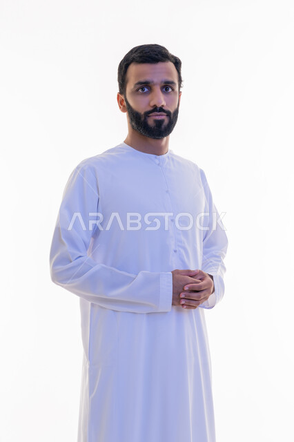 مفهوم الرجولة والقوة والثقة بالنفس، النظر إلى الكاميرا بايماءات الجدية والسرور، الاهتمام بالأناقة والمظهر الخارجي، بورتريه لرجل عربي خليجي اماراتي يرتدي الكندورة يقف بأيدي متشابكة بتعابير الحزم، خلفية بيضاء