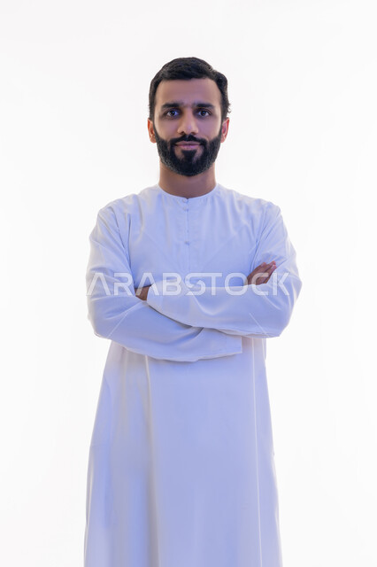 النظر إلى الكاميرا بايماءات الجدية والسرور، الاهتمام بالأناقة والمظهر الخارجي، مفهوم الرجولة والقوة والثقة بالنفس، بورتريه لرجل عربي خليجي اماراتي يرتدي الكندورة يقف بأيدي متقاطعة بتعابير الحزم، خلفية بيضاء
