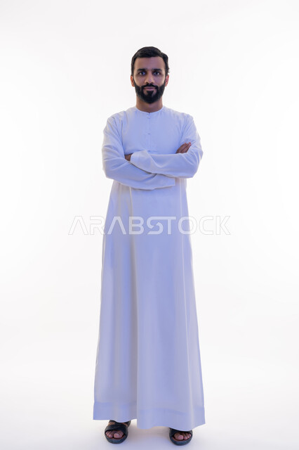 الاهتمام بالأناقة والمظهر الخارجي، النظر إلى الكاميرا بايماءات الجدية والسرور، مفهوم الرجولة والقوة والثقة بالنفس، بورتريه لرجل عربي خليجي اماراتي يرتدي الكندورة يقف بأيدي متقاطعة بتعابير الحزم، الطول الكامل للجسم، خلفية بيضاء