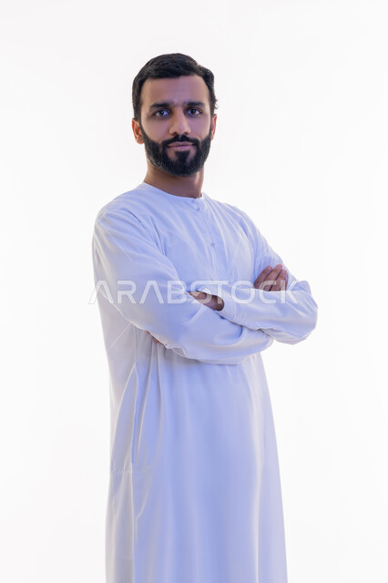 النظر إلى الكاميرا بايماءات الجدية والسرور، الاهتمام بالأناقة والمظهر الخارجي، مفهوم الرجولة والقوة والثقة بالنفس، بورتريه لرجل عربي خليجي اماراتي يرتدي الكندورة يقف بأيدي متقاطعة بتعابير الحزم، خلفية بيضاء