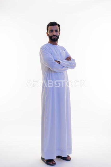 الاهتمام بالأناقة والمظهر الخارجي، النظر إلى الكاميرا بايماءات الجدية والسرور، مفهوم الرجولة والقوة والثقة بالنفس، بورتريه لرجل عربي خليجي اماراتي يرتدي الكندورة يقف بأيدي متقاطعة بتعابير الحزم، الطول الكامل للجسم، خلفية بيضاء