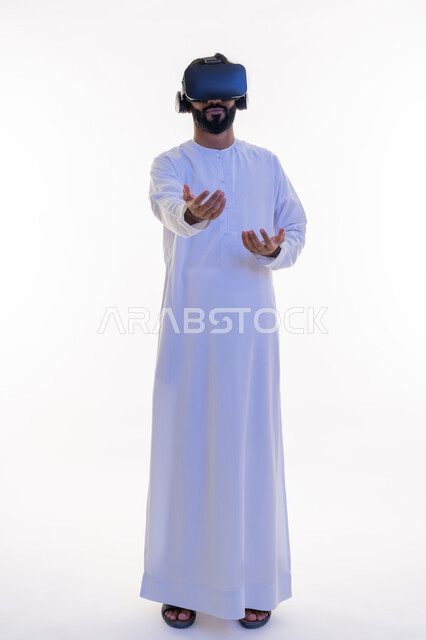 رفع اليدين بإيماءات الاندماج والاستمتاع، مشاهدة الأفلام باستخدام تقنيات الواقع الافتراضي, محاكاة العالم الخيالي, بورتريه لرجل عربي خليجي إماراتي يرتدي الكندورة يضع نظارة ثلاثية الأبعاد يقوم بالتحرك والتفاعل معها، الجسم بالطول الكامل، خلفية بيضاء