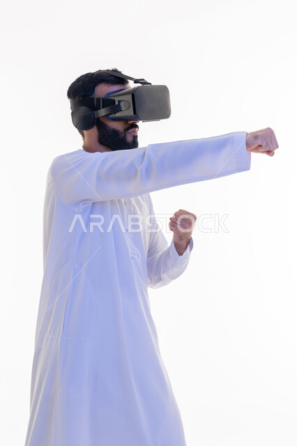 ممارسة رياضة الدفاع عن النفس بتقنيات تكنولوجية، بورتريه من الجانب لرجل عربي خليجي إماراتي يرتدي الكندورة ونظارات الواقع الافتراضي يرفع يديه بإيماءات الملاكمة والمصارعة، التدرب على الحركات القتالية من خلال ألعاب الفيديو، خلفية بيضاء