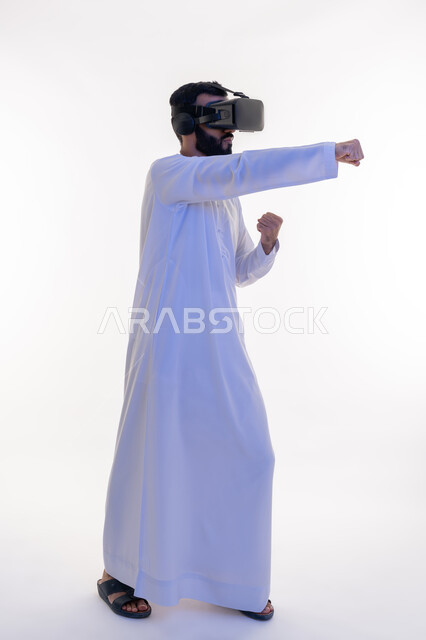 التدرب على الحركات القتالية من خلال ألعاب الفيديو، ممارسة رياضة الدفاع عن النفس بتقنيات تكنولوجية، بورتريه من الجانب لرجل عربي خليجي إماراتي يرتدي الكندورة ونظارات الواقع الافتراضي يرفع يديه بإيماءات الملاكمة والمصارعة، الجسم بالطول الكامل، خلفية بيضاء