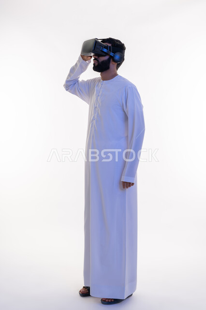 محاكاة العالم الخيالي، الاستمتاع بالمشاهدة عبر نظارة الواقع الافتراضي، بورتريه لرجل عربي خليجي إماراتي يرتدي الكندورة ونظارات ثلاثية الأبعاد يضع يديه عليها، إيماءات الاندماج والتركيز، استخدام الأجهزة التقنية الحديثة، الجسم بالطول الكامل، خلفية بيضاء