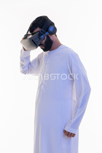 Gestures of integration and concentration, use of modern technical devices, enjoyment and entertainment by watching through virtual reality glasses, portrait of an Arab Gulf Emirati man wearing a kandura looking through 3D glasses with his hand on them, integration and simulation of the imaginary world, white background