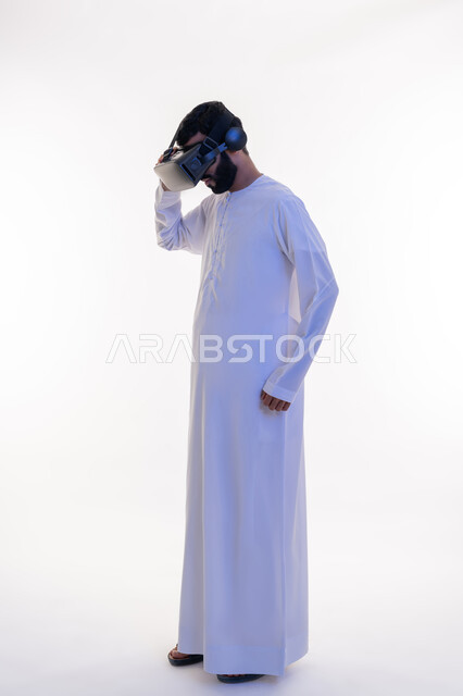 محاكاة العالم الخيالي، الاستمتاع بالمشاهدة عبر نظارة الواقع الافتراضي، بورتريه لرجل عربي خليجي إماراتي يرتدي الكندورة ونظارات ثلاثية الأبعاد يضع يديه عليها، إيماءات الاندماج والتركيز، استخدام الأجهزة التقنية الحديثة، الجسم بالطول الكامل، خلفية بيضاء