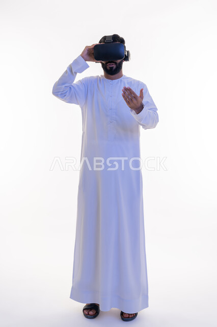 الاستمتاع بالمشاهدة عبر نظارة الواقع الافتراضي، بورتريه لرجل عربي خليجي إماراتي يرتدي الكندورة ونظارات ثلاثية الأبعاد يرفع يده للأعلى، محاكاة العالم الخيالي، إيماءات الاندماج والتركيز، استخدام الأجهزة التقنية الحديثة، الجسم بالطول الكامل، خلفية بيضاء