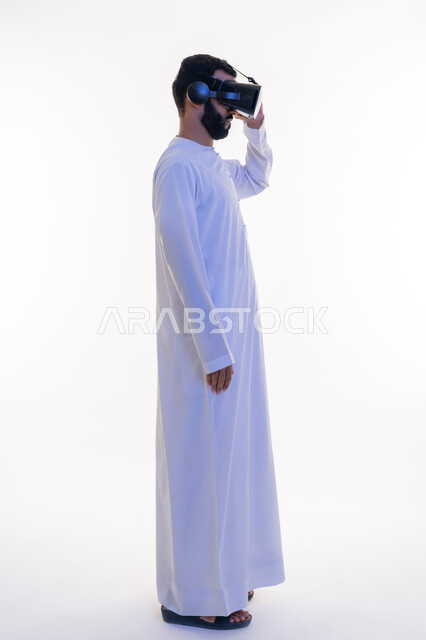 الاستمتاع بالمشاهدة عبر نظارة الواقع الافتراضي، بورتريه من الجانب لرجل عربي خليجي إماراتي يرتدي الكندورة ونظارات ثلاثية الأبعاد يضع يده عليها، إيماءات الاندماج والتركيز، استخدام الأجهزة التقنية الحديثة، محاكاة العالم الخيالي، الجسم بالطول الكامل، خلفية بيضاء