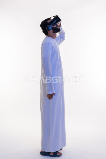 استخدام الأجهزة التقنية الحديثة، محاكاة العالم الخيالي، الاستمتاع بالمشاهدة عبر نظارة الواقع الافتراضي، بورتريه من الجانب لرجل عربي خليجي إماراتي يرتدي الكندورة ونظارات ثلاثية الأبعاد يضع يده عليها، إيماءات الاندماج والتركيز، الجسم بالطول الكامل، خلفية بيضاء
