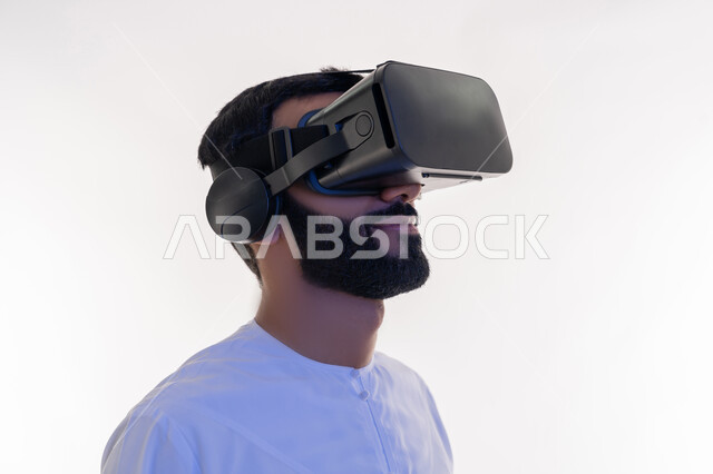 المتعة والتسلية بالمشاهدة عبر نظارة الواقع الافتراضي، بورتريه مقرب لرجل عربي خليجي إماراتي يرتدي الكندورة ينظر من خلال نظارات ثلاثية الأبعاد، الاندماج ومحاكاة العالم الخيالي، ايماءات السعادة والسرور، التطور التقني والتكنولوجي, خلفيه بيضاء